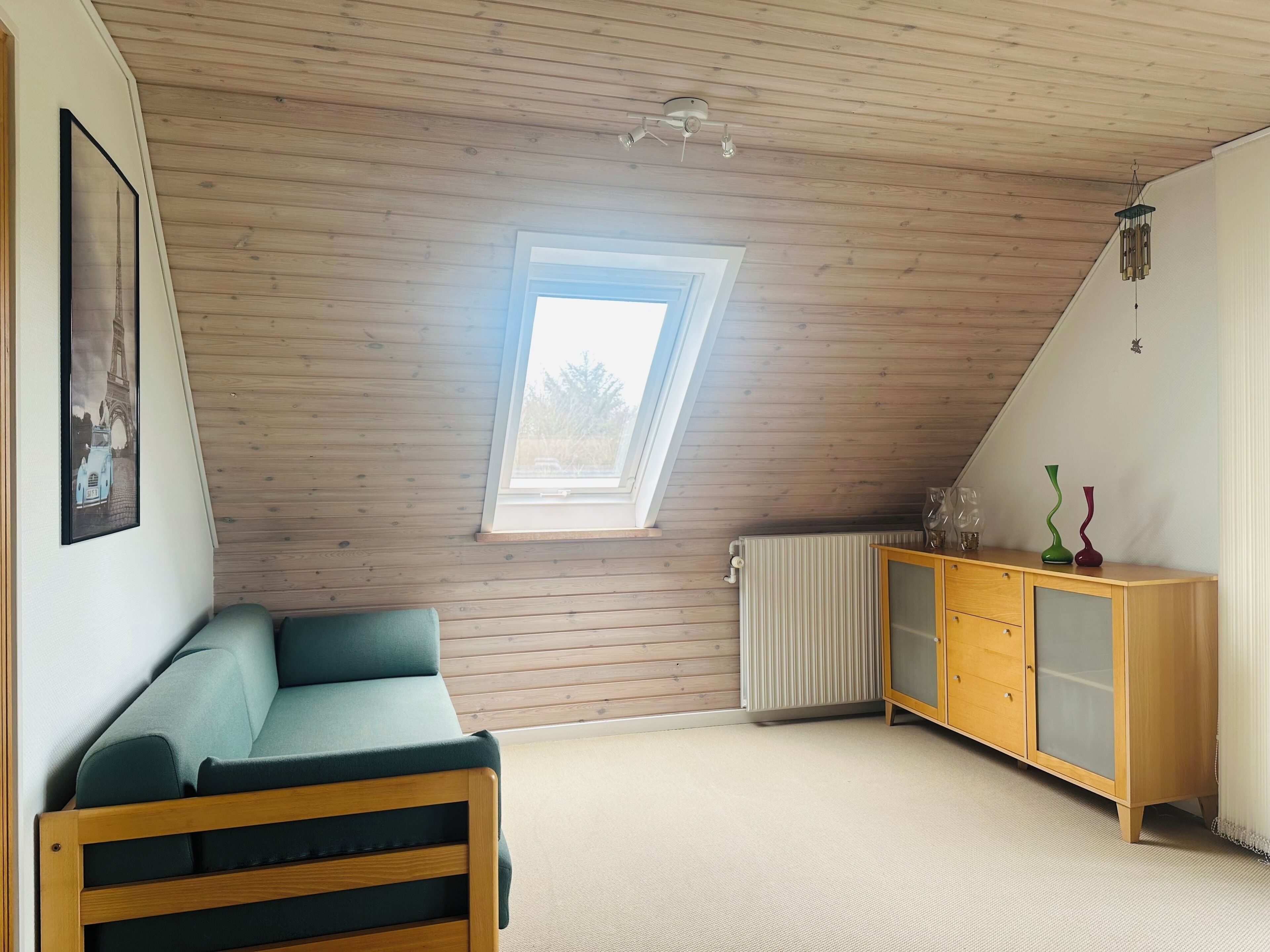 A room with a sloped ceiling, a window, a green sofa, a wooden cabinet, and a radiator.