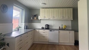 A kitchen with white cabinets, a clock, a window with blinds, and a plant. - Family private house 4-5 rooms (Brovst)