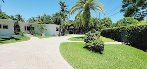 A paved driveway leading to a white building with a covered patio area, surrounded by lush greenery and palm trees.