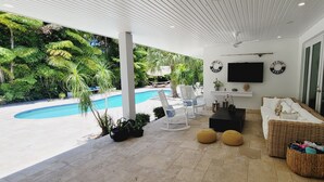 Ett takförsett utomhusområde med sittplatser, en pool, vita möbler och en platt-TV.