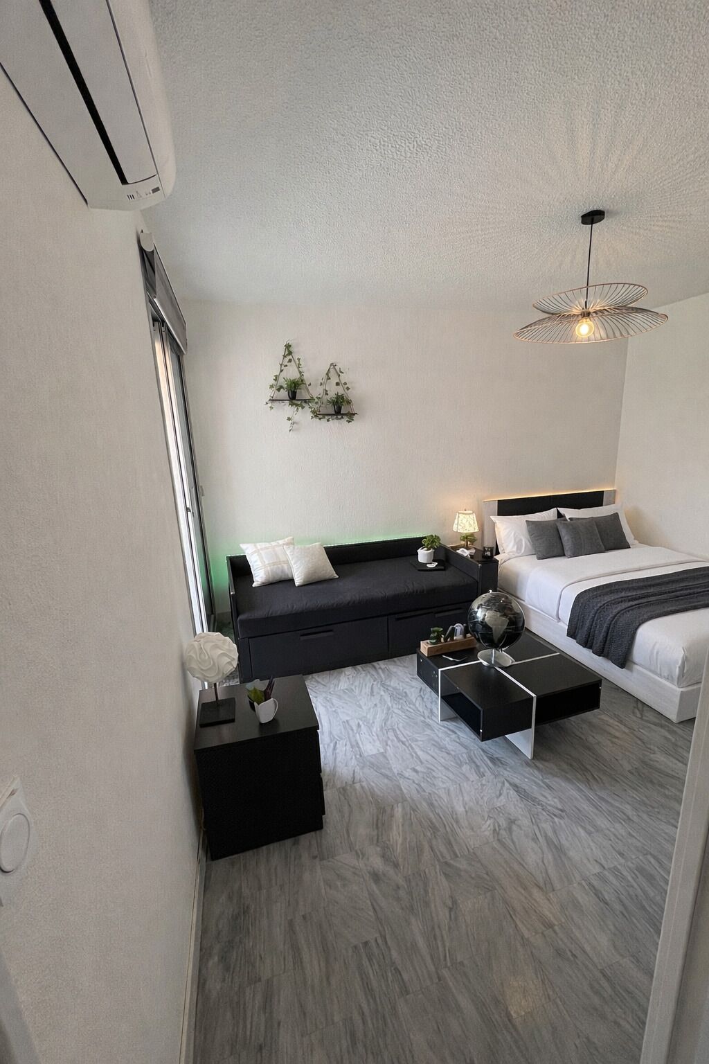 A modern bedroom with a bed, a sofa, a coffee table, a hanging light fixture, and a wall-mounted shelf with plants.
