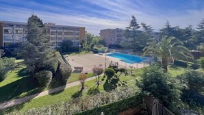 A pool surrounded by greenery, a building with multiple floors, and a clear sky. - Apartment with swimming pool, janitor and parking space (Mandelieu-la-Napoule)