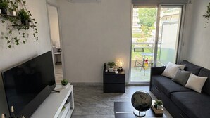 A modern living room with a flat-screen TV, a black sofa, a coffee table, and a hanging light fixture. - Apartment with swimming pool, janitor and parking space (Mandelieu-la-Napoule)