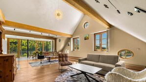 Ein geräumiges Wohnzimmer mit hohen Decken, Holzbalken, einem großen Fenster, einer Couch, einem Sessel und einem Tisch.