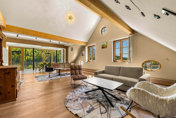 A spacious living room with a high ceiling, wooden beams, a large window, a sofa, a chair, and a table.