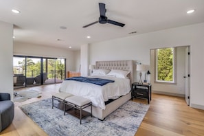 Une chambre avec un grand lit, un ventilateur de plafond, une table de chevet, un banc et une vue sur l’extérieur, accessible par une porte coulissante.
