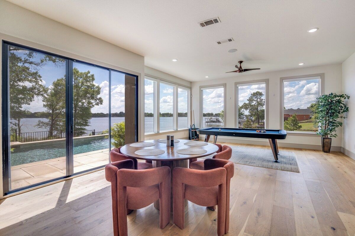 Une pièce spacieuse équipée d’une table de billard, d’une table ronde avec des chaises et de grandes fenêtres donnant sur un lac.