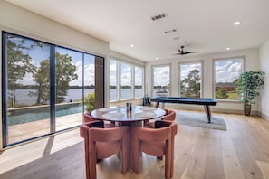 Een ruime kamer met een pooltafel, een ronde tafel met stoelen en grote ramen met uitzicht op een meer.