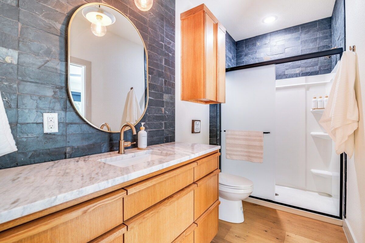 A bathroom with a marble countertop, wooden cabinets, a large mirror, a toilet, and a glass shower enclosure.