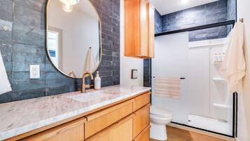 A bathroom with a marble countertop, wooden cabinets, a large mirror, a toilet, and a glass shower enclosure.