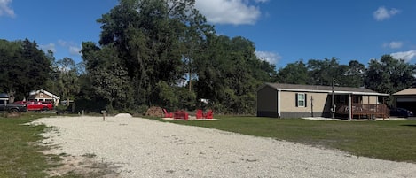 A residential area with a large tree, a gravel driveway, a house with a porch, and a red vehicle parked nearby.