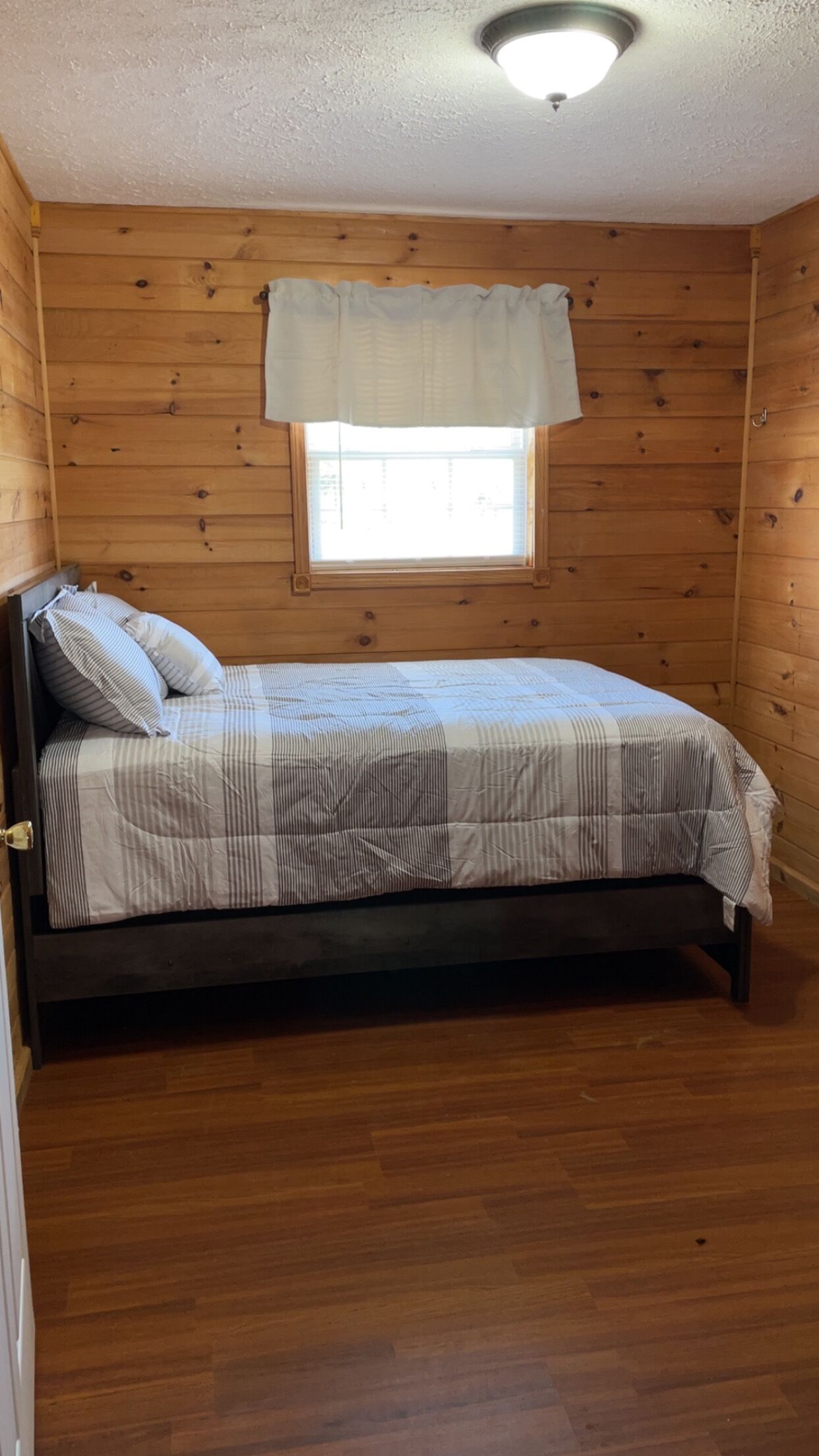 Ein Zimmer mit Holzwänden, einem Bett, einem Fenster mit weißem Vorhang und einer Deckenleuchte.