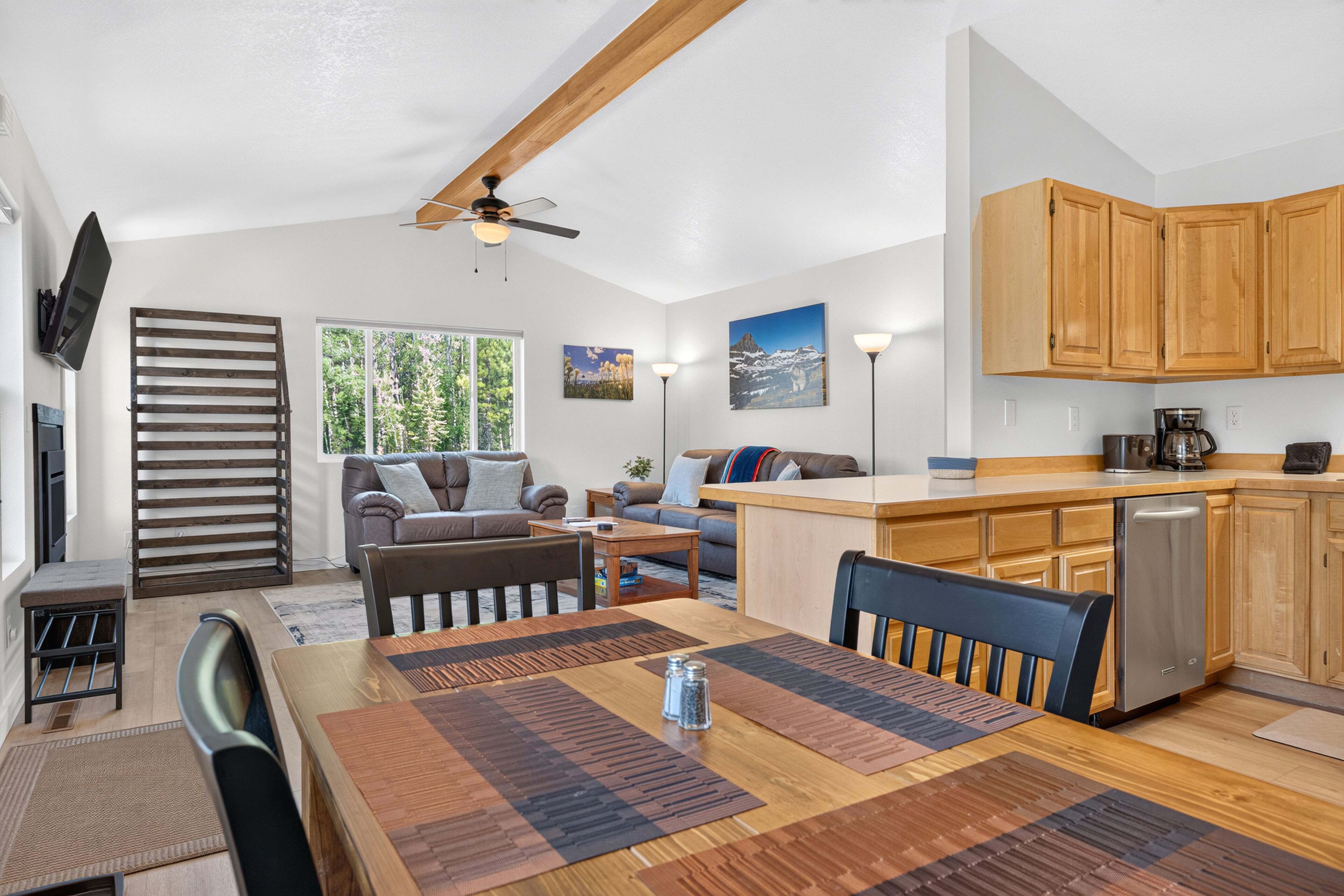 A modern kitchen with wooden cabinets, a dining area with a wooden table and chairs, a sofa, and a TV mounted on the wall.
