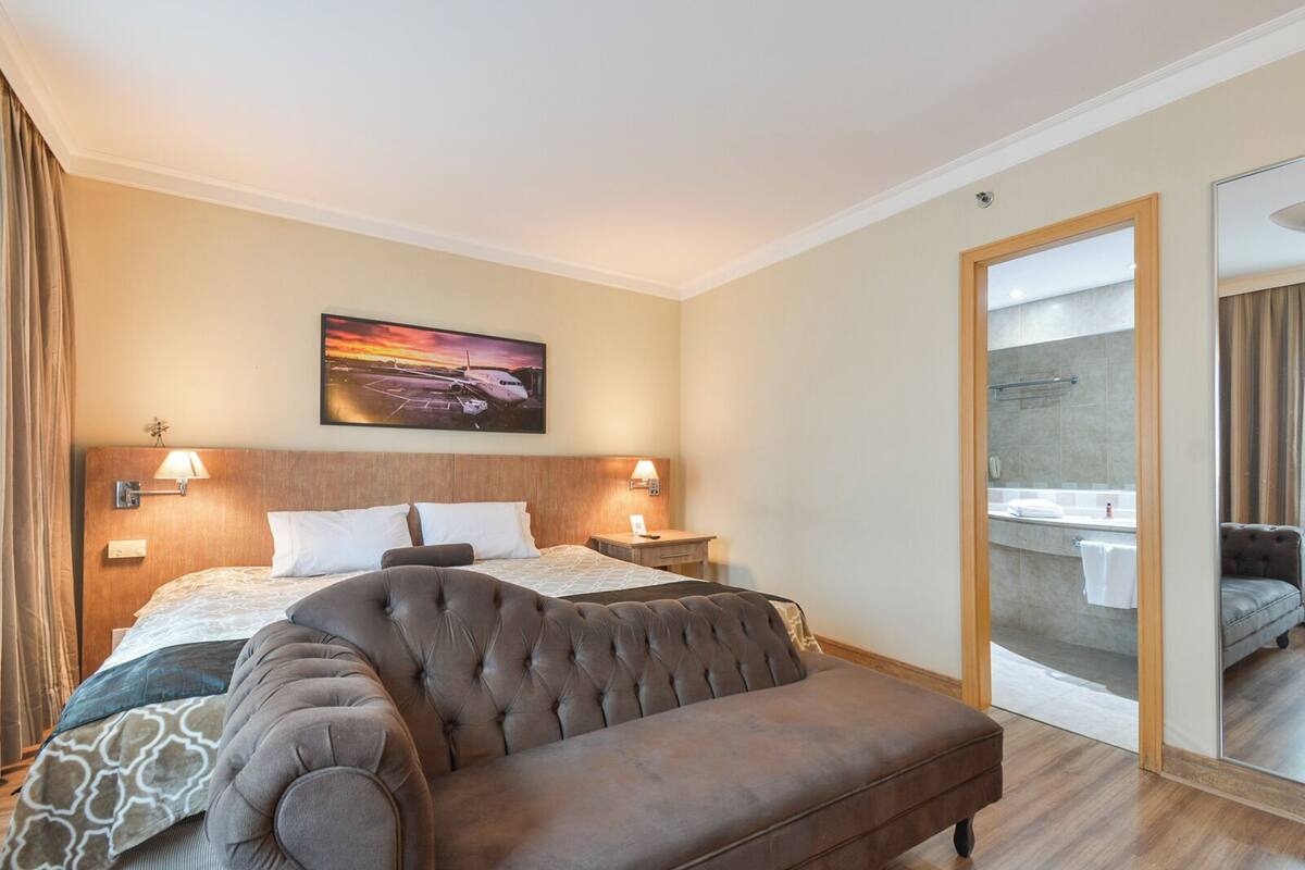 A hotel room with a large bed, two bedside lamps, a headboard, a large window, and a bathroom visible through an open door.