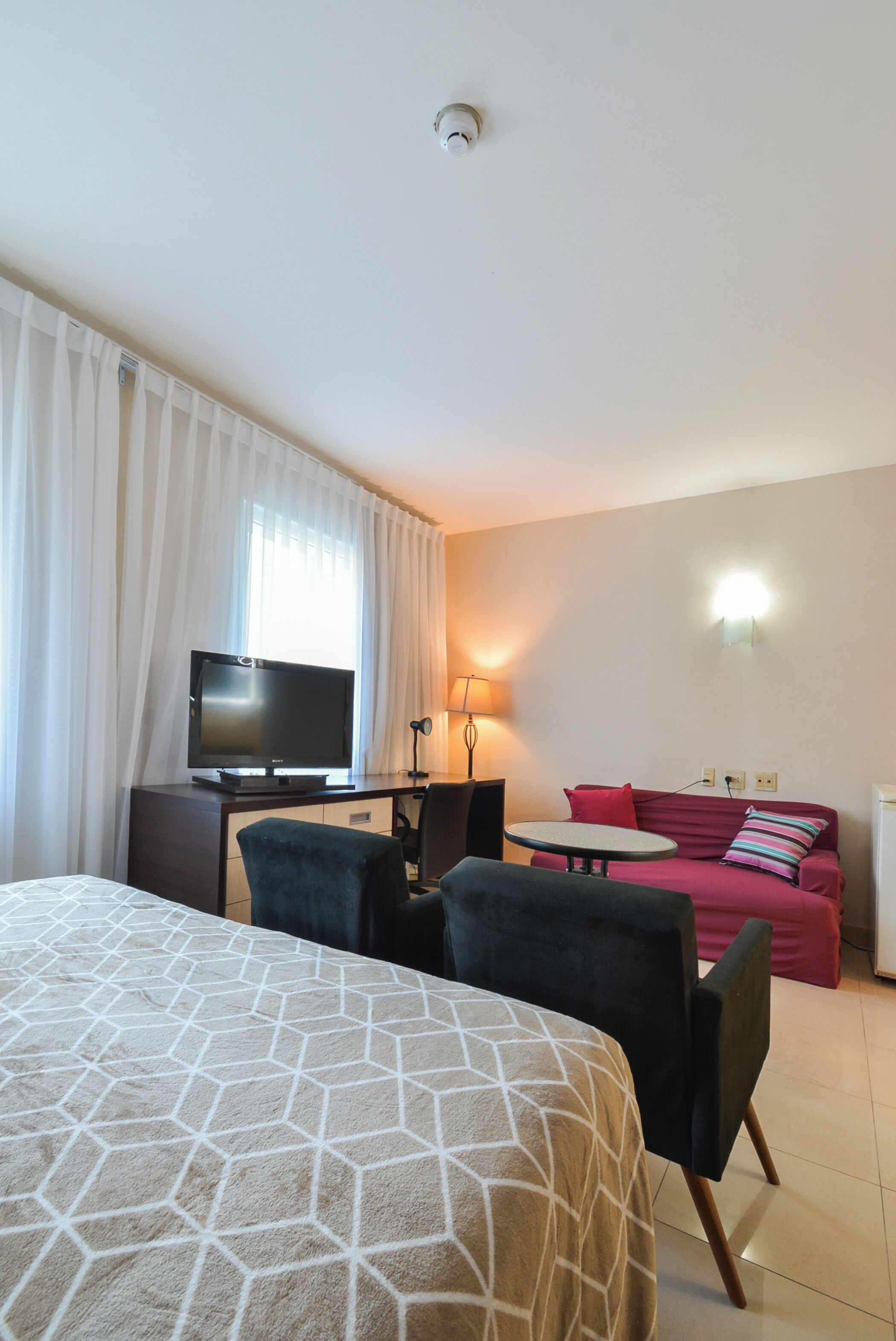 A hotel room with a bed, a desk with a television, a chair, a small table, a sofa, and a lamp.