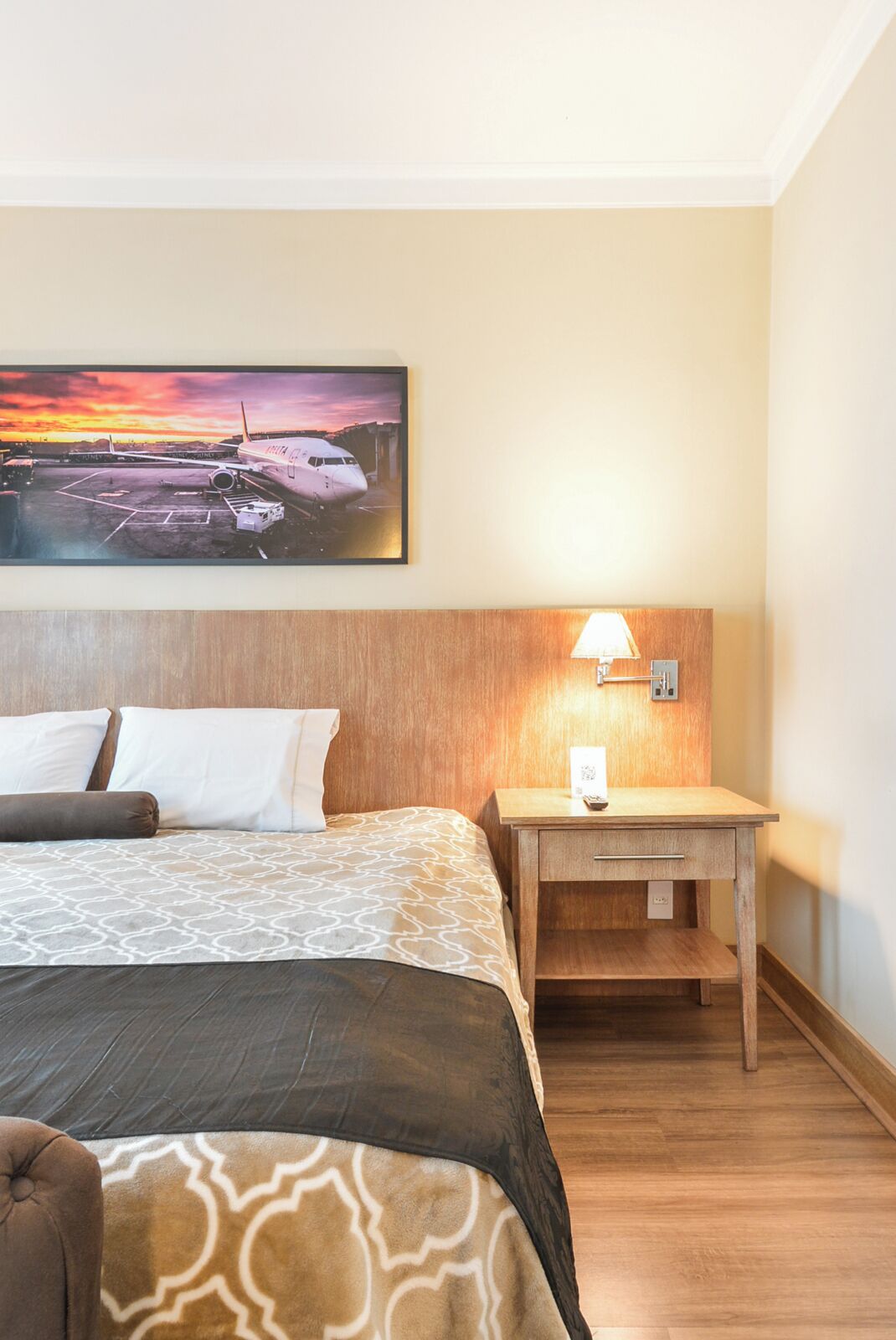 A hotel room with a bed, a desk, a nightstand, a lamp, and a painting of an airplane on the wall.