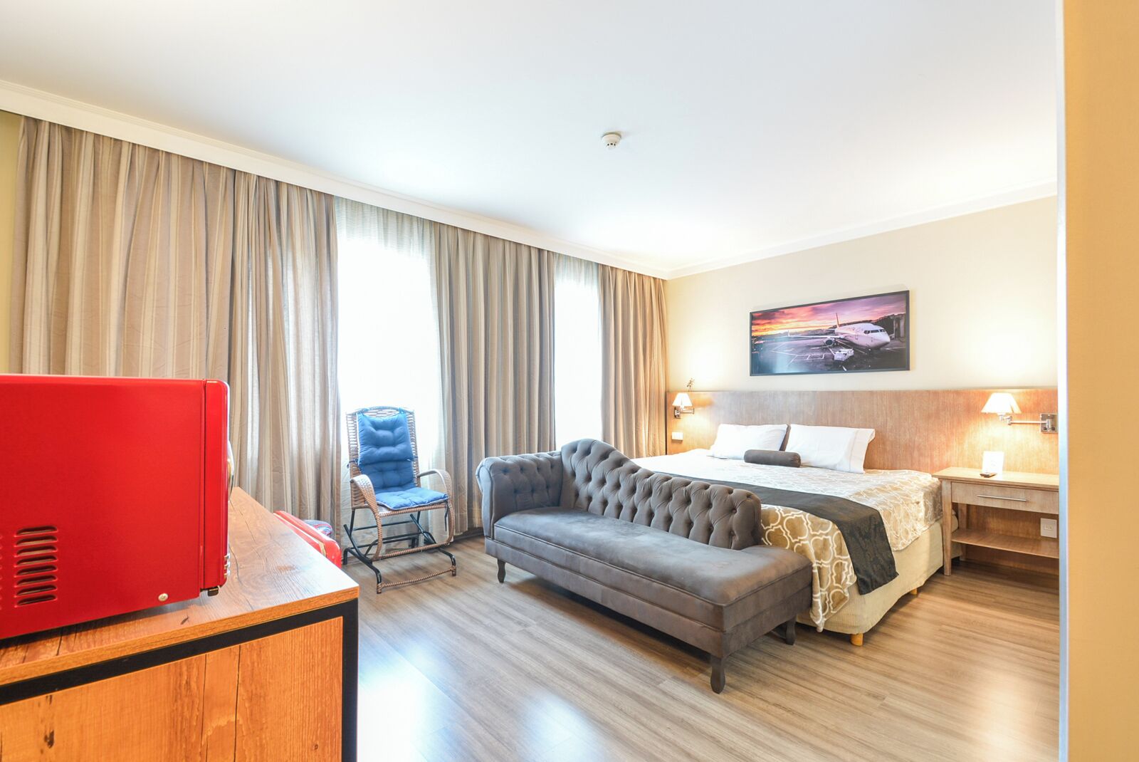 A hotel room with a bed, a grey sofa, a red desk, a wicker chair, and a painting on the wall.
