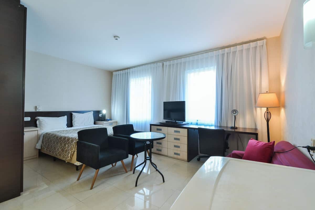 A hotel room with a bed, a desk, a chair, a small table, a TV, and a lamp.