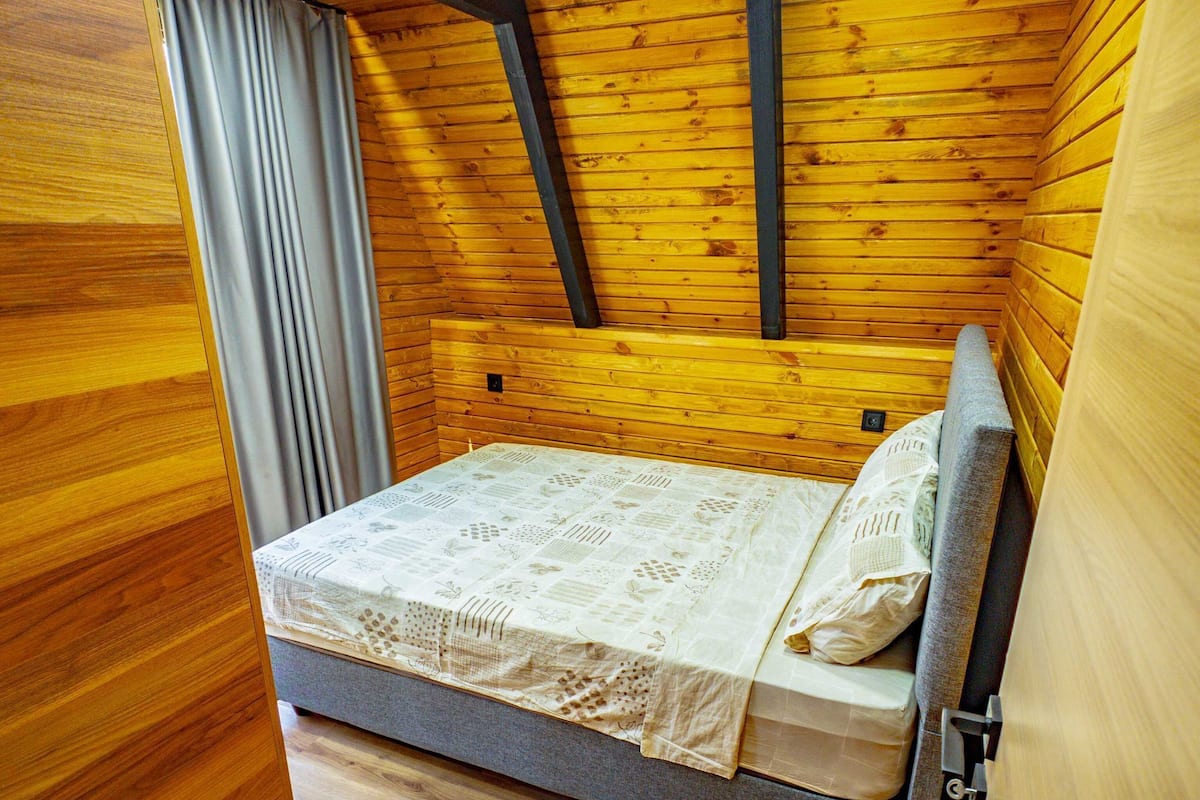 A small wooden room with a bed, a window with curtains, and a sloped ceiling.