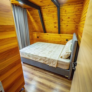 A small wooden room with a bed, a window with curtains, and a sloped ceiling.