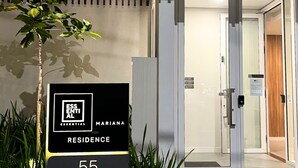 Entrada moderna de un edificio con puerta de vidrio, un cartel que indica 'ESSENTIAL MARINA RESIDENCE 55' y un pequeño árbol en primer plano.