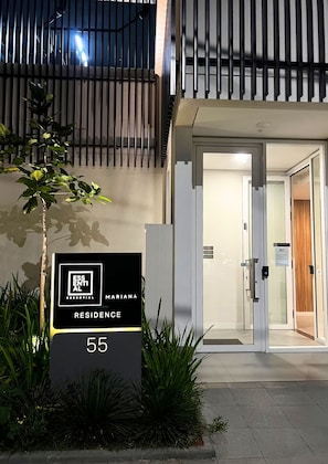 Entrada de um edifício moderno com porta de vidro, sinal indicando 'ESSENTIAL MARINA RESIDENCE 55' e uma pequena árvore em primeiro plano.