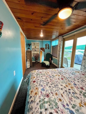Un dormitorio con colcha estampada, techo de madera, vistas a un lago y una silla junto a la ventana.