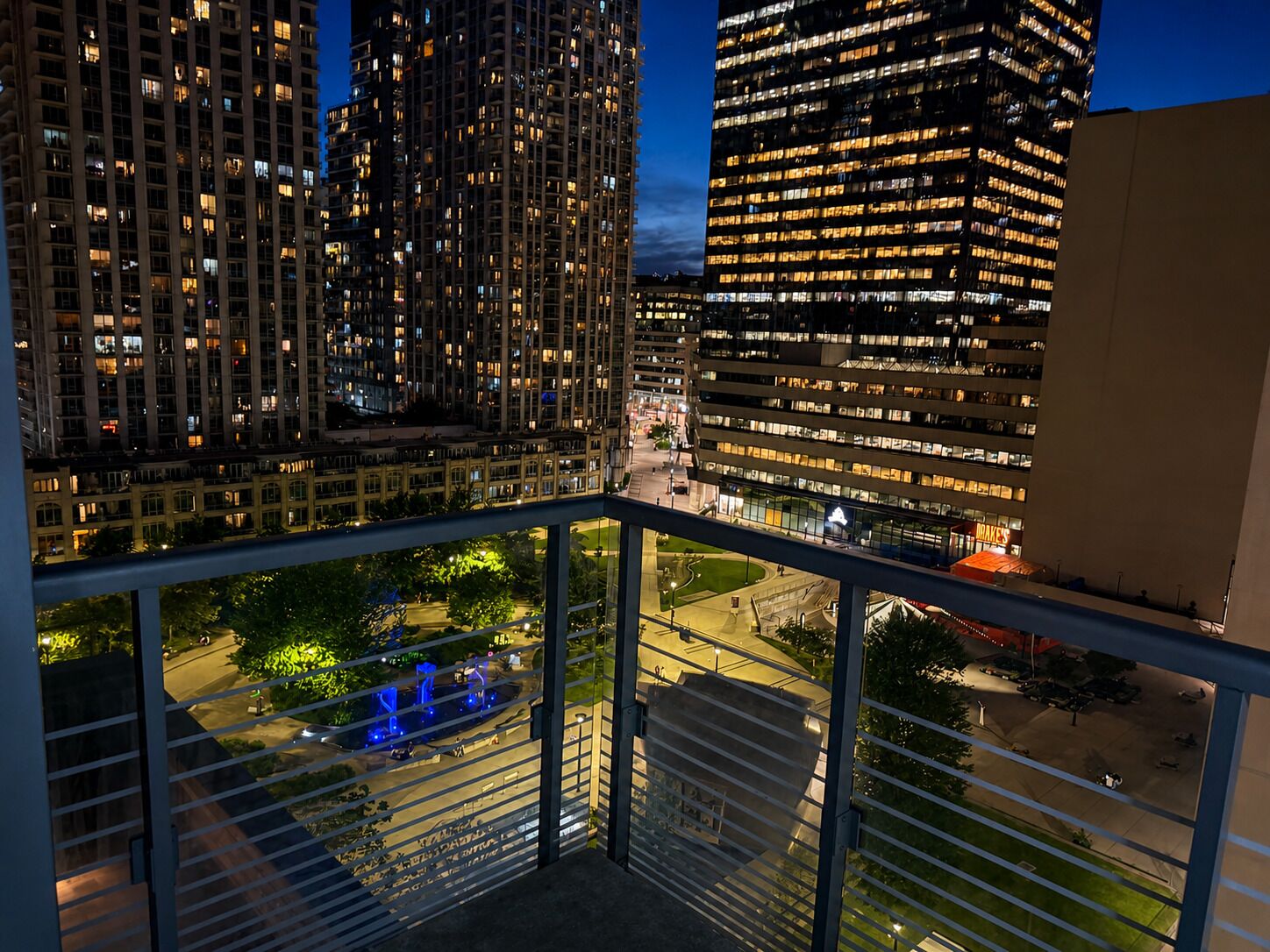 Une vue nocturne d’une ville avec de hauts immeubles, une place bien éclairée ornée de sculptures et une vue dégagée sur l’environnement urbain.