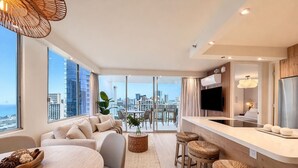 A modern living room with a sofa, dining table, and bar stools. There is a large window with a city view. - Waikiki Ocean Luxe Retreat | Panoramic Views (Honolulu)