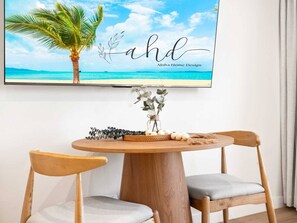 Ein Essbereich mit einem runden Holztisch, zwei Holzstühlen, einer Vase mit Grünpflanzen und einem an der Wand montierten Fernseher, der eine tropische Strandlandschaft zeigt.