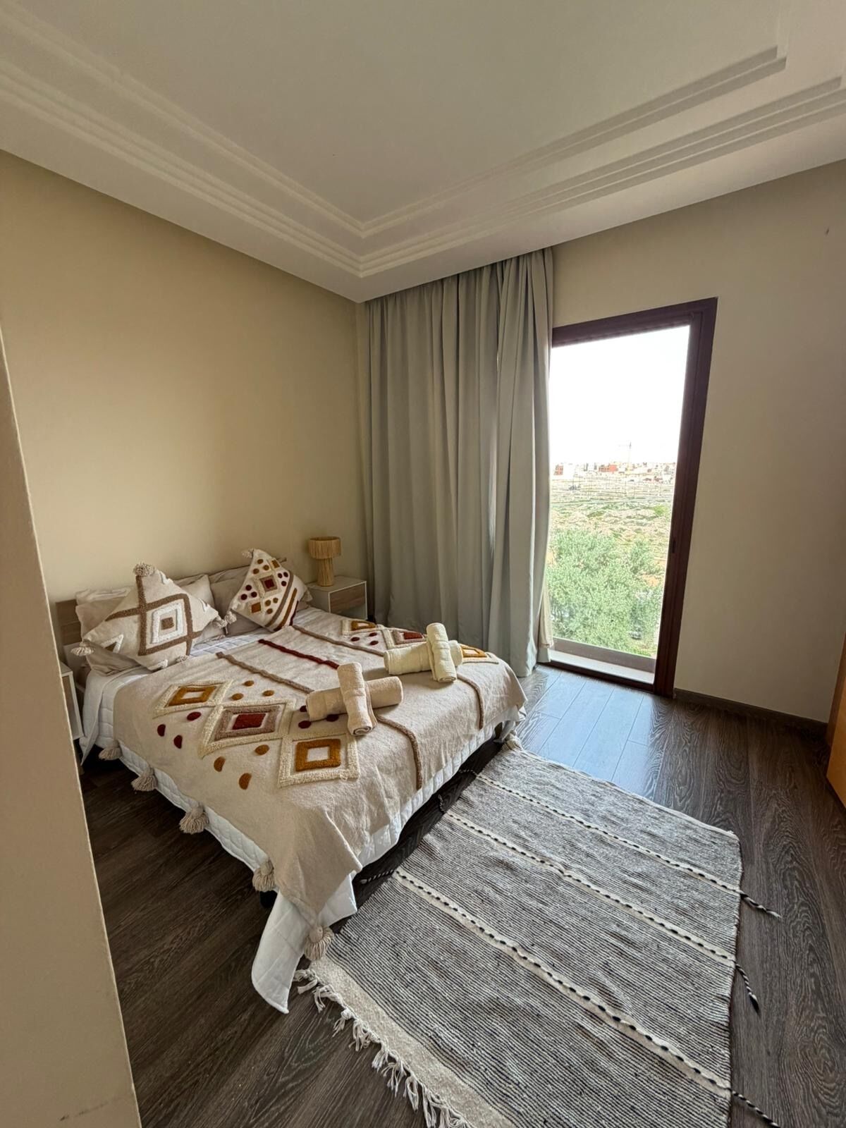 Un dormitorio con cama, mesita de noche, lámpara y vistas al verdor exterior.