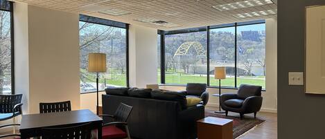 Una sala de estar moderna con un sofá, sillas, una mesa de comedor y grandes ventanales que ofrecen vistas a un puente y un parque.