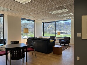 Una sala de estar moderna con un sofá, sillas, una mesa de comedor y grandes ventanales que ofrecen vistas a un puente y un parque.