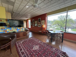 A spacious living area with a large window, a flat-screen TV, a dining table, and a patterned rug.