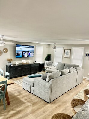 A living room with a grey sofa, a TV, a dark wooden cabinet, and a dining area with a table and chairs. -  Steps away to the beach - Surf - Sun & Sand beach house (Hampton)