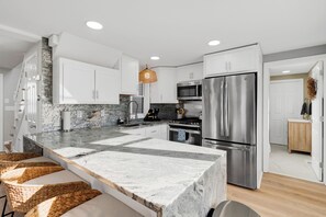 A modern kitchen with white cabinets, a granite countertop, stainless steel appliances, and a hanging light fixture. -  Steps away to the beach - Surf - Sun & Sand beach house (Hampton)