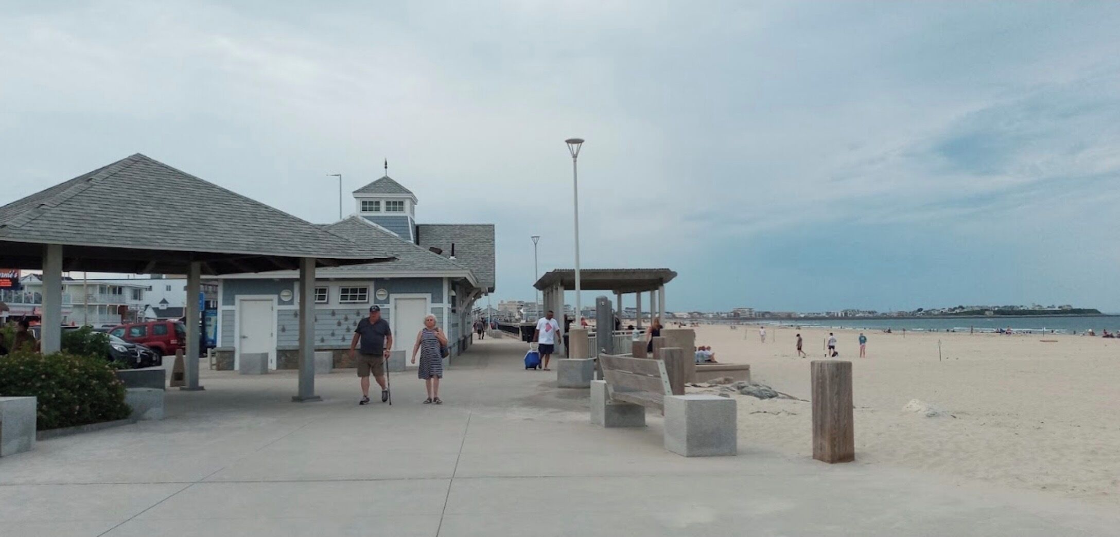Un paseo marítimo en la playa con un área techada, un muelle y una playa de arena con gente.