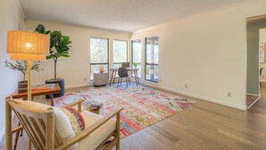 Une pièce spacieuse dotée d’un plancher en bois, d’une grande fenêtre, d’une chaise en bois, d’une table avec une lampe et d’un tapis coloré.