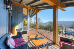 Una terraza techada al aire libre con mobiliario de mimbre, una mesa de madera y vistas a las montañas.