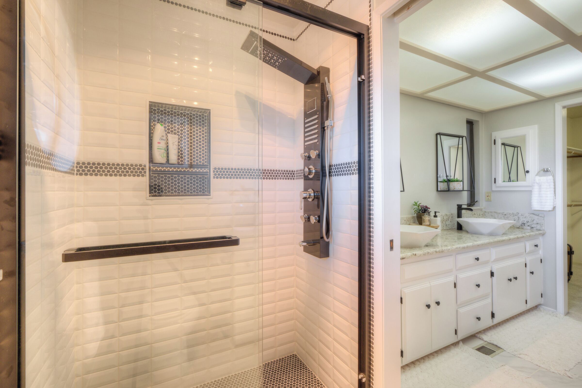 A modern bathroom with a glass shower enclosure, a white tiled wall, a sink with a marble countertop, and a mirror above the sink.