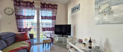 Un salon avec un canapé, un téléviseur, une table basse en verre sur laquelle sont posés des verres à vin, et un tableau accroché au mur.