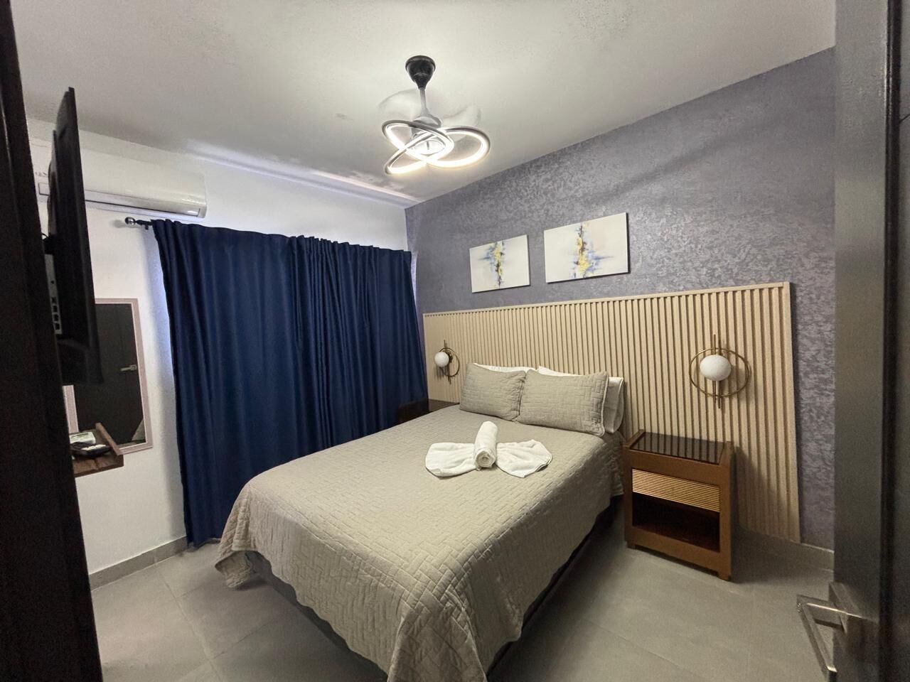 A hotel room with a bed, a bedside table, a wall-mounted lamp, and a ceiling fan.