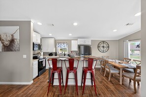 A modern kitchen with white cabinets, a central island with red bar stools, a dining table with wooden chairs, and a large clock on the wall.