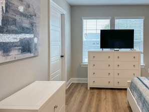 A bedroom with a dresser, a bed, a television on a stand, and a window with blinds.