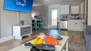 Een woonkamer met een flatscreen televisie, een salontafel met pingpong rackets, gele en oranje ballen, een klok, en een keuken met witte kasten en een koelkast.