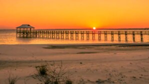 Een strand met een pier die in het water reikt, een klein gebouwtje aan het einde van de pier en de zon die ondergaat op de achtergrond.