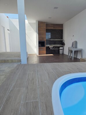 A modern outdoor living area with a pool, a kitchen with dark marble backsplash, and a seating area with plastic chairs.