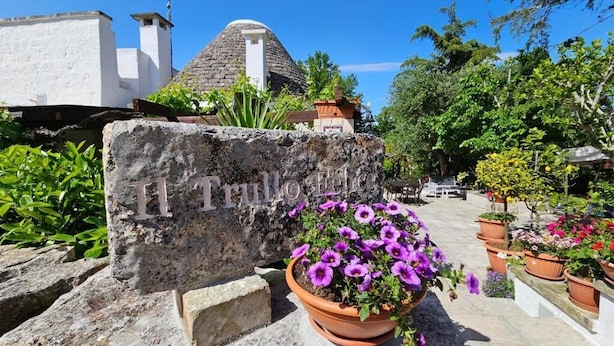 En stenskilt med teksten 'Il Trullo', en stenhvælvet bygning og en potteplante med lilla blomster.
