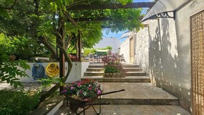 En stentrappe fører op til et overdækket område med et pergola, en trillebør med blomster og en haveslange.