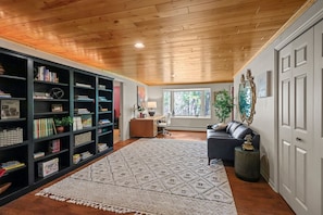 Una habitación con techo de madera, una estantería, un escritorio, una silla, un sofá, una alfombra y una ventana.